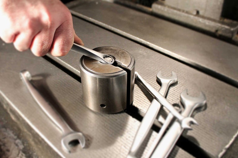En hand använder en skiftnyckel på en metallcylinder, omgiven av andra skiftnycklar på en arbetsbänk.