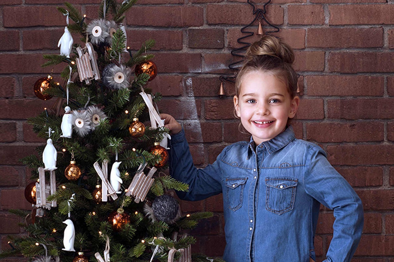 Flicka bredvid en julgran med julgransdekorationer som kulor, pingviner och träslädar framför en tegelvägg