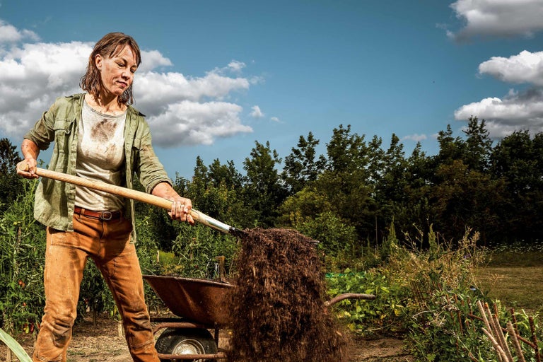 Kvinna skyfflar jord med en spade i en skottkärra i trädgården.