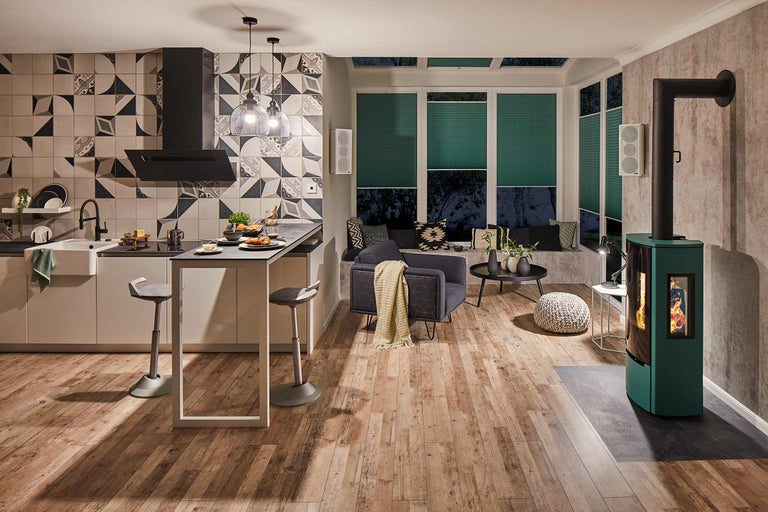 Ljust vardagsrum med kök, sittgrupp vid fönstret, vedspis och trägolv.