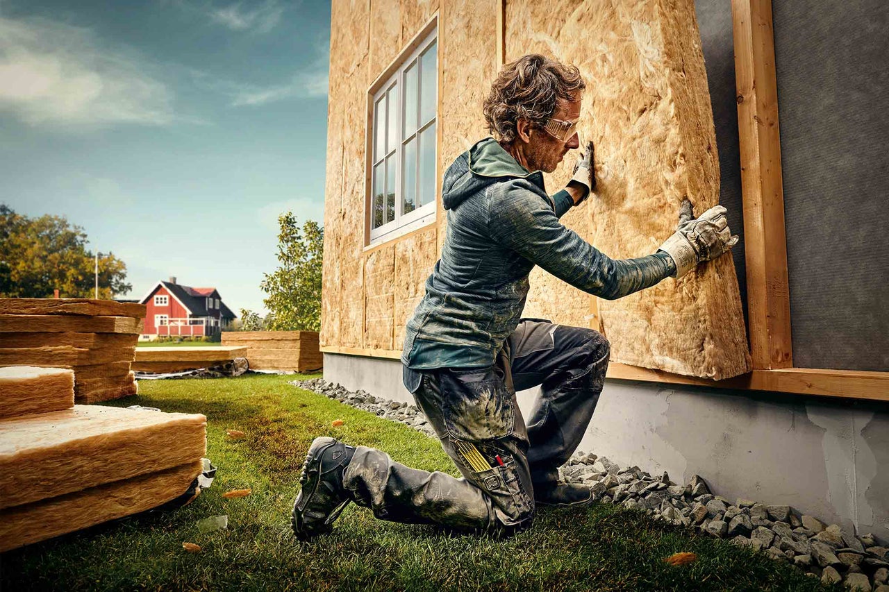 En man knäböjer framför en husvägg och fäster isoleringsull, med staplad isoleringsull och ett rött hus i bakgrunden.