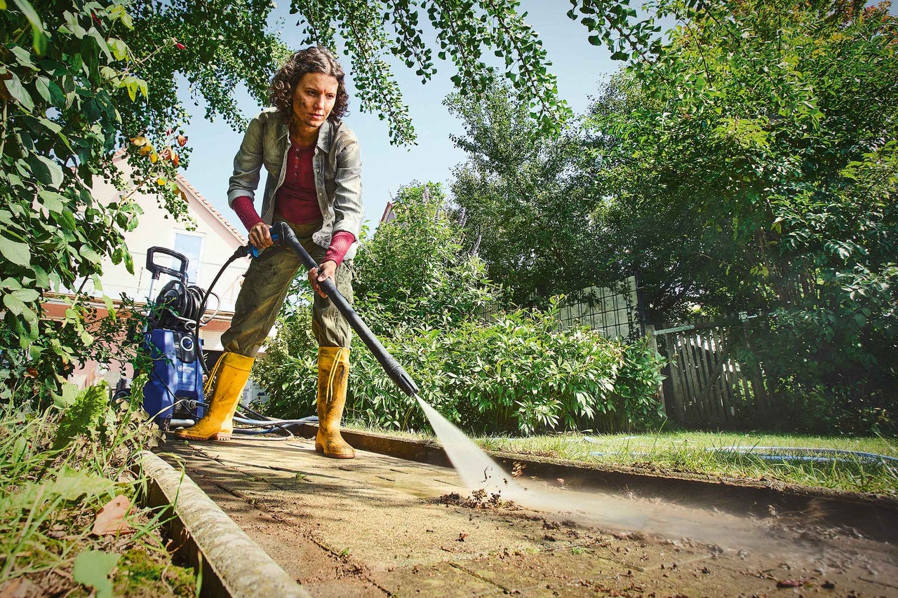 Kvinna rengör gångväg med högtryckstvätt i trädgården.