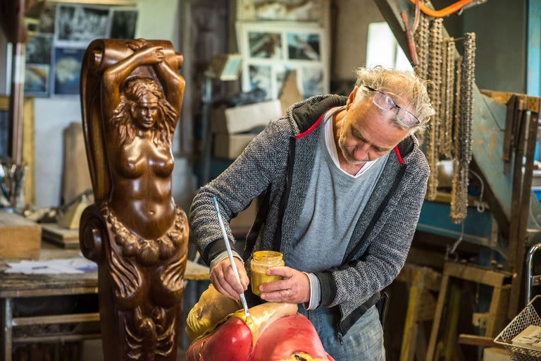 En konstnär målar en skulptur i en verkstad. I bakgrunden står en träskulptur av en kvinna.