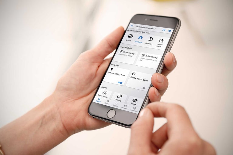 En person använder en smart hem app på en smartphone för att styra belysning, solskydd och olika scenarier.