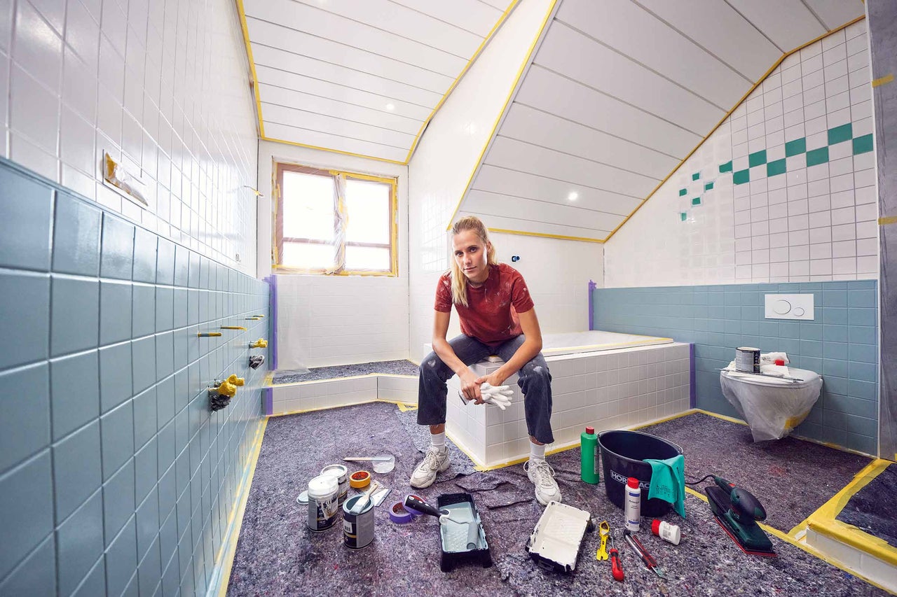 En kvinna sitter i ett badrum som håller på att renoveras, omgiven av färgburkar och målarverktyg på ett skyddande underlag.