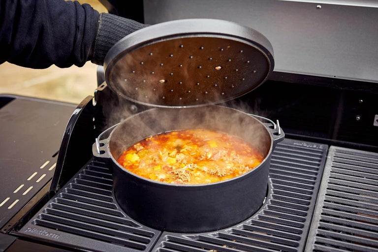 En holländsk ugn med lock står på en grill, fylld med en maträtt från vilken ånga stiger.