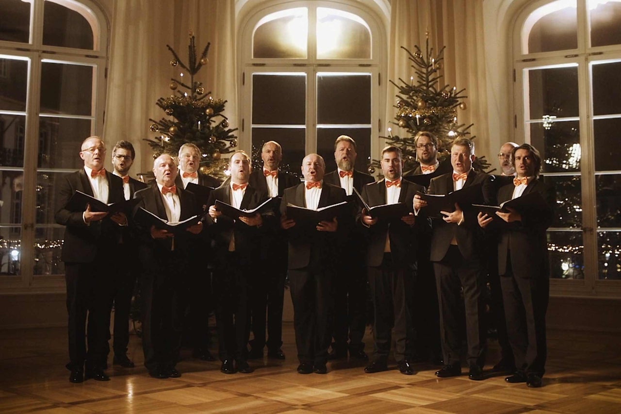 En manskör i kostymer och flugor sjunger framför fönster och julgranar.