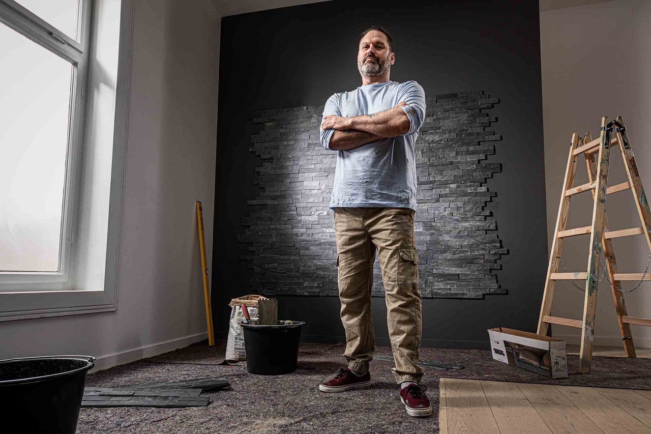 En man står med armarna i kors i ett rum med väggbeklädnad av stenpaneler, verktyg och en stege.