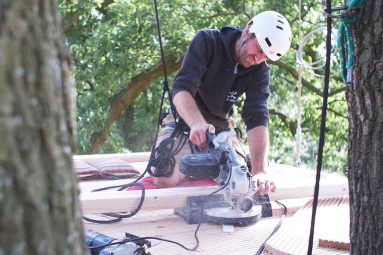 Man med skyddshjälm sågar trä med en kap- och gersåg under byggandet av ett trädkoja.