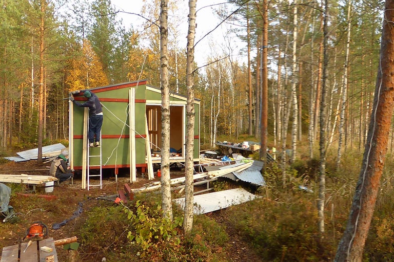 Ett trähus byggs i skogen. En person arbetar på en stege på husets fasad.