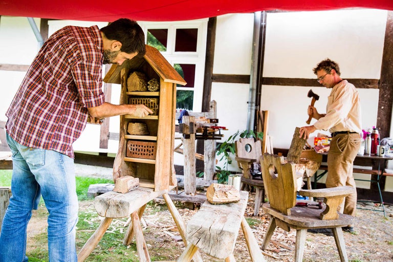 Man fyller ett insektshotell med bomaterial, medan en annan man hugger ved i bakgrunden