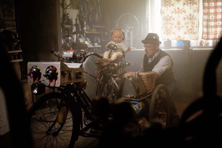 En man med hatt och glasögon står bredvid en modifierad cykel med en korg och en docka som förare i en verkstad.