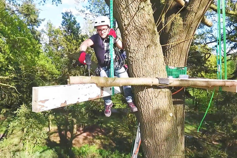 Man med hjälm och säkerhetsutrustning arbetar med borrmaskin på trädplattform