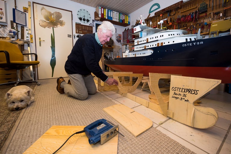Man arbetar på en fartygsmodell med en sticksåg i en verkstad, med en hund i bakgrunden