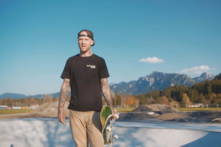 En man med tatueringar håller en skateboard i en skatepark.