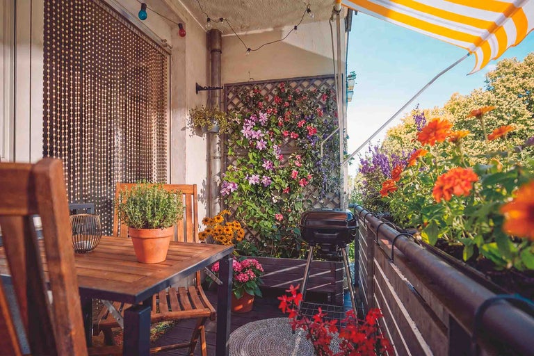 Mysig balkong med trämöbler, klotgrill, blommor i krukor, spaljé med klätterväxter och gul-vitrandig markis.
