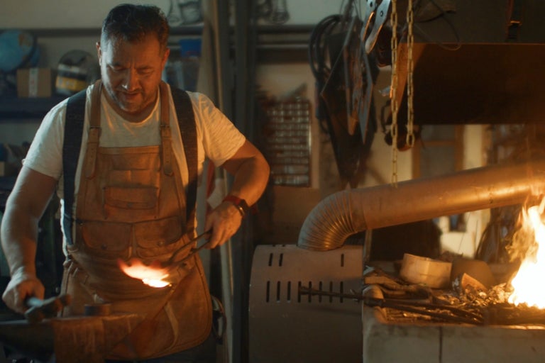 En man med förkläde av läder smider metall i en verkstad med en smedja
