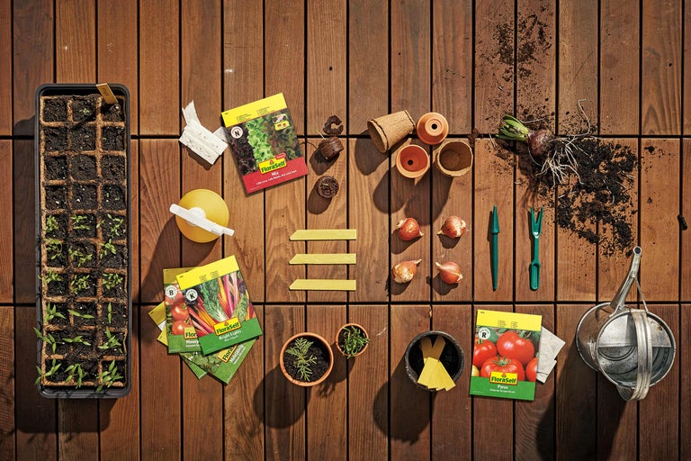 Trädgårdsscen med odlingslåda, fröpåsar från FloraSelf, blomkrukor och trädgårdsredskap på trägolv.