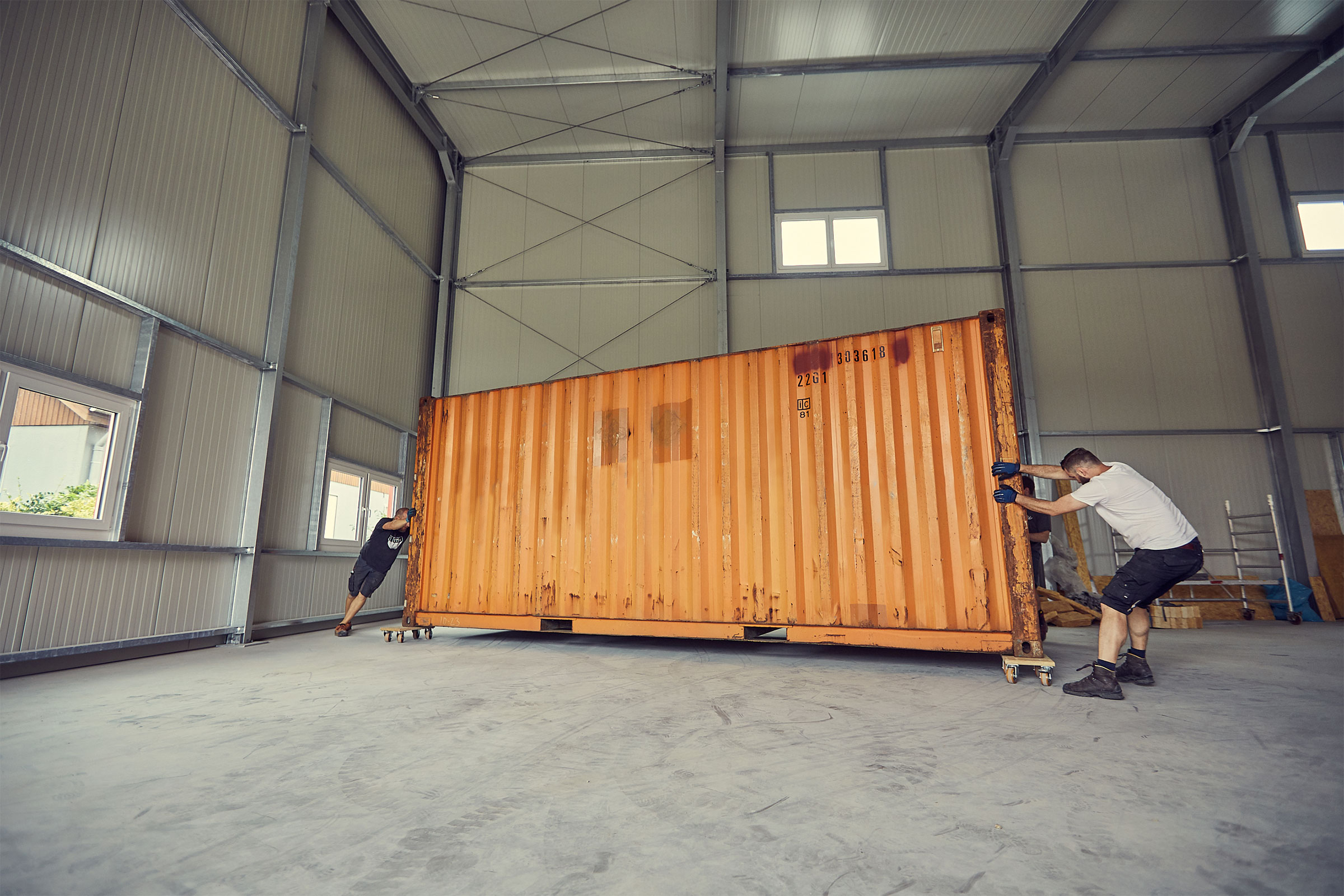 Två personer flyttar en förvaringscontainer på hjul i ett lager med metallstruktur.
