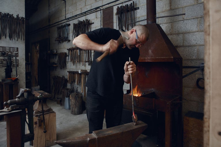 En smed bearbetar glödande metall med en hammare på ett städ i sin verkstad.