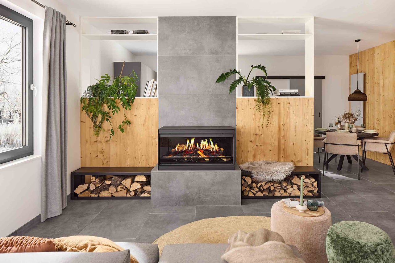 Vardagsrum med öppen spis, vedförvaring, växter och sittplatser för en mysig atmosfär.