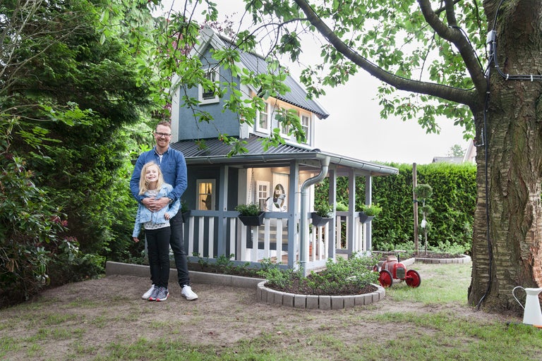 En man kramar en flicka framför ett blått lekstuga med ett grått metalltak i trädgården.