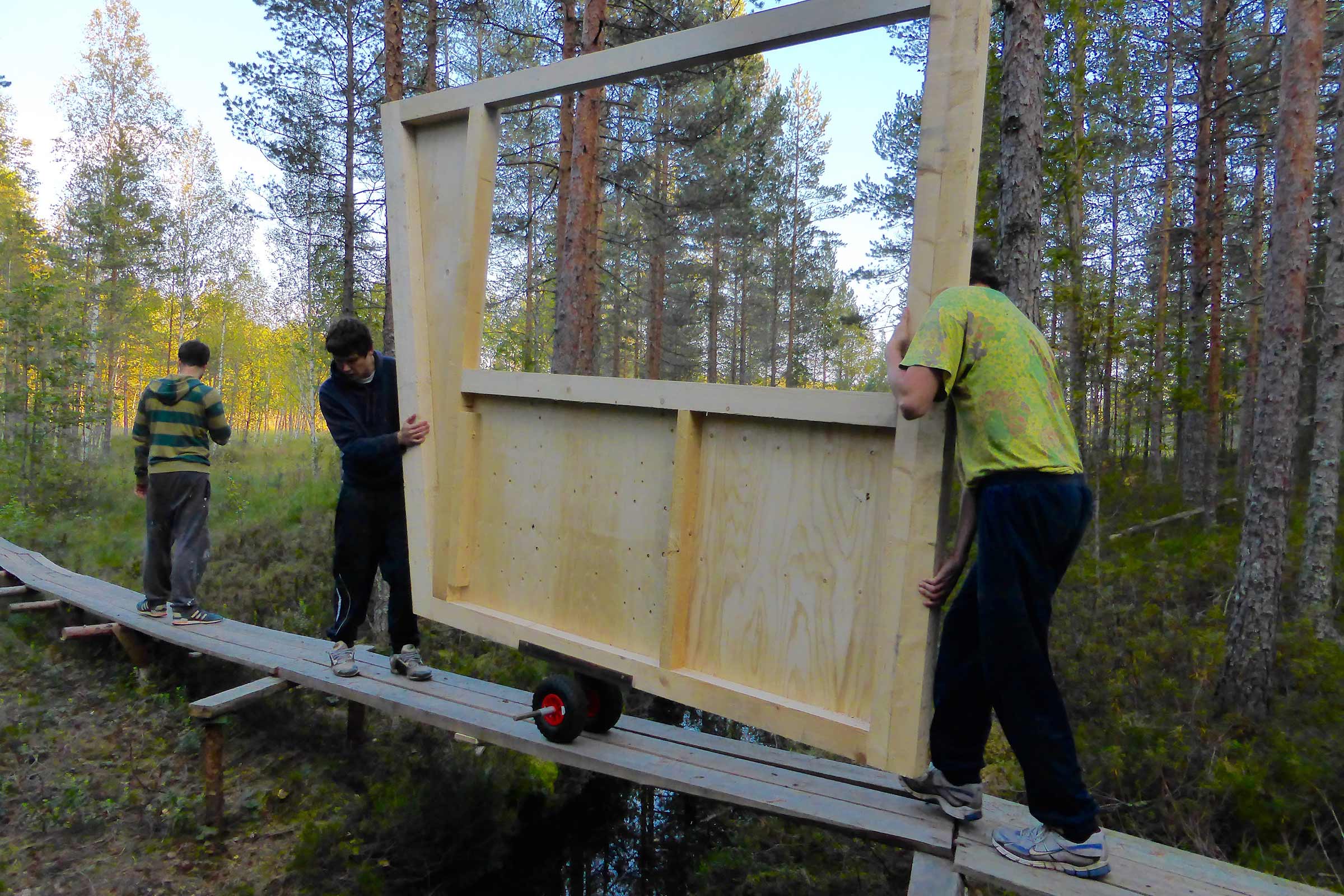 Två personer transporterar en träramkonstruktion med hjul över en träbro.