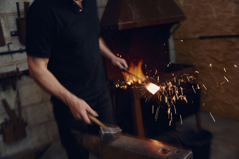 Smed som bearbetar glödande metall med en hammare på ett städ i smedjan