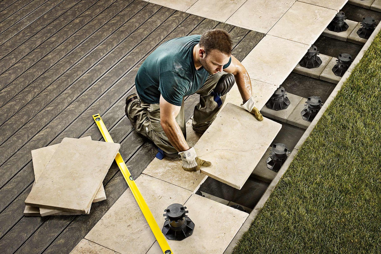 En man lägger stenplattor på en terrass med justerbara stöd och ett vattenpass.