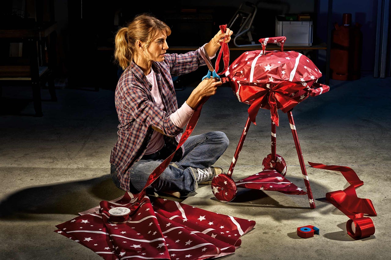 En kvinna sitter på golvet och klipper ett band bredvid en grill insvept i papper och en rosett.