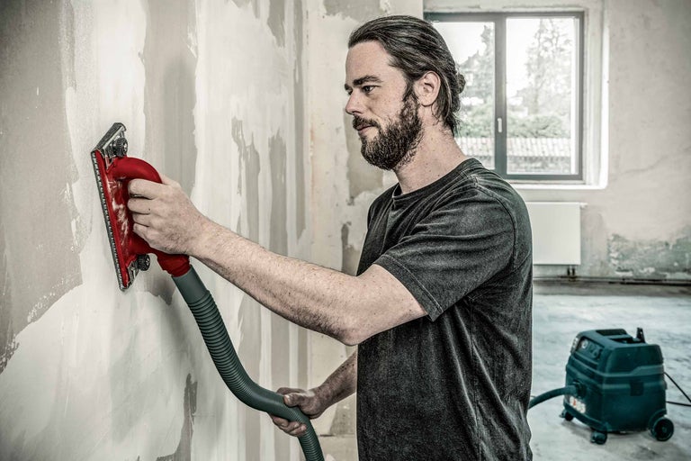 Man slipar vägg med en gipsskiveslipmaskin, ansluten till en dammsugare