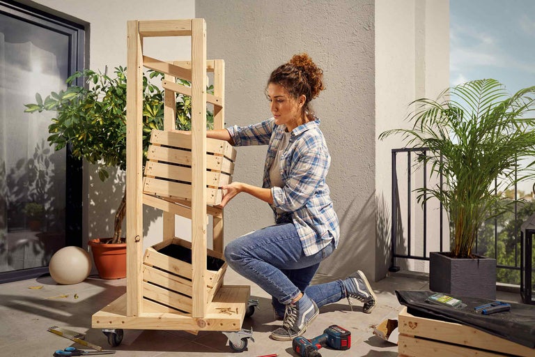 Kvinna monterar en mobil träplanteringslåda på en balkong med verktyg och växter.