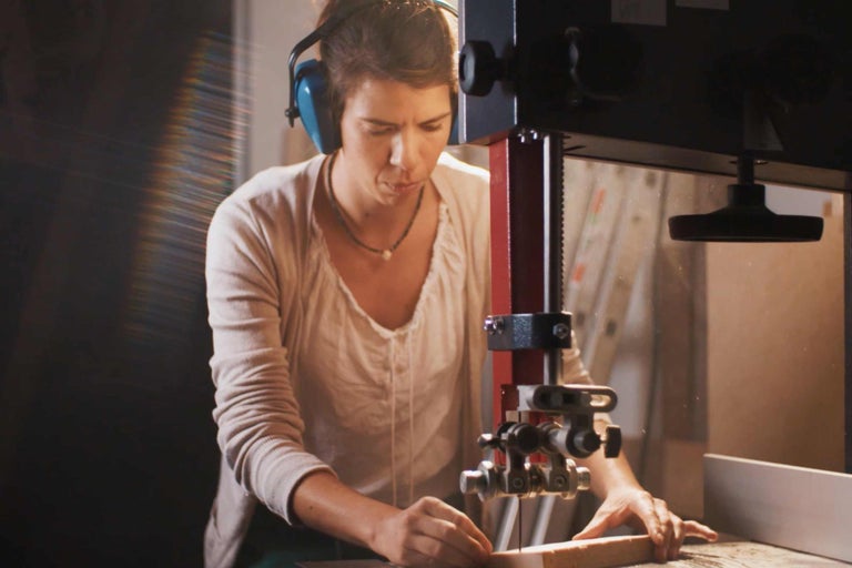 En kvinna sågar en bit trä med en bandsåg medan hon bär hörselskydd.