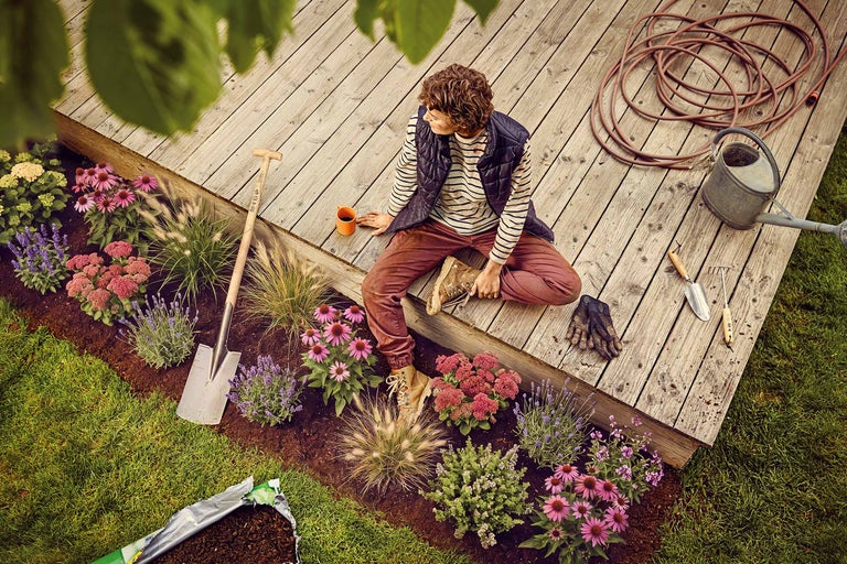 Kvinna som sitter på en träterrass bredvid en rabatt med spade, vattenkanna, trädgårdsslang och olika blommor som solhatt och lavendel.