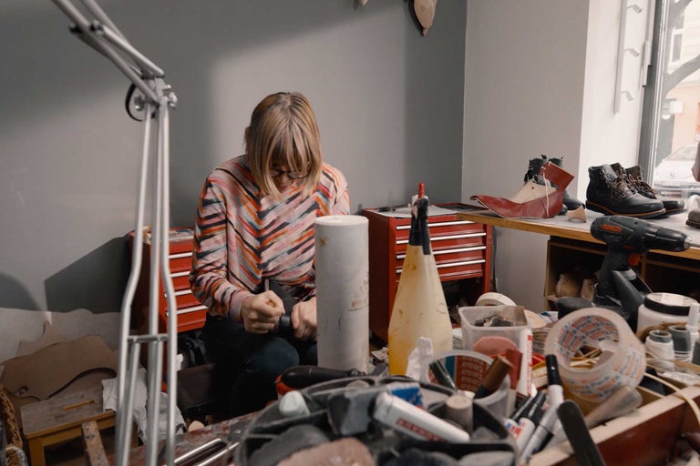 En kvinna arbetar med att tillverka skor i sin verkstad, verktyg och material ligger på bordet.