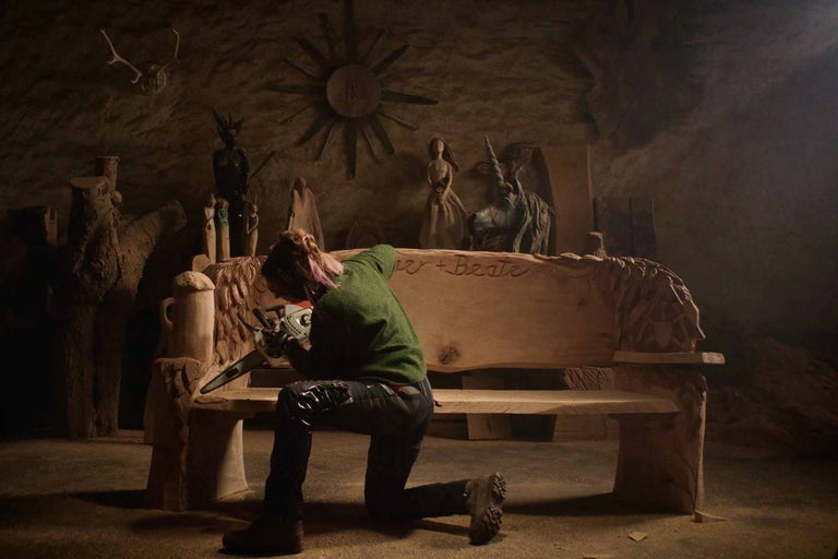 En kvinna knäböjer framför en träbänk och arbetar på den med en motorsåg. I bakgrunden finns olika träskulpturer.