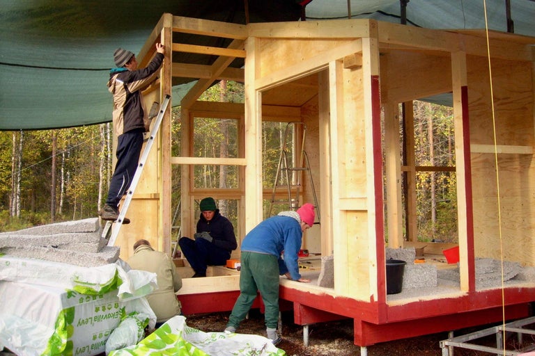 Flera personer bygger ett trähus med träram och isoleringsskivor. Byggarbetsplatsen ligger utomhus.