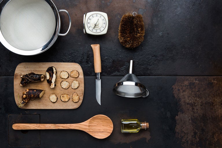 Köksredskap och ingredienser på mörk bakgrund, inklusive sil, timer, borste, kniv, tratt, olja och träbräda med rotgrönsaker.
