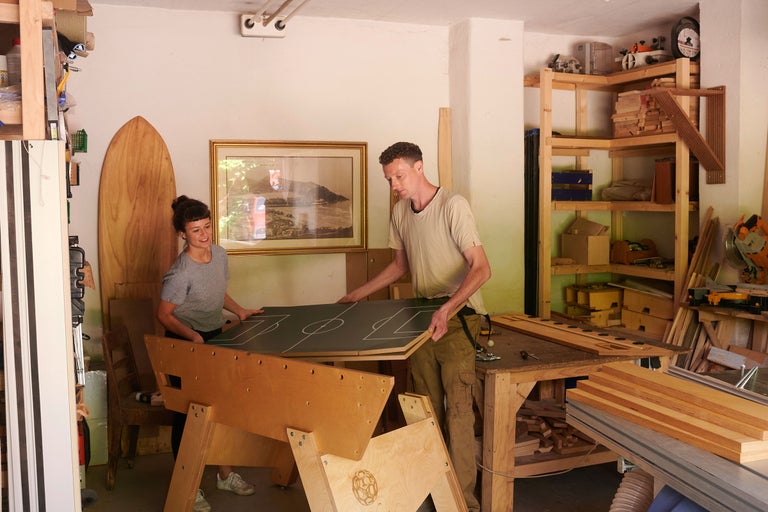 Två personer monterar ett fotbollsspel i en verkstad med trähyllor och verktyg.