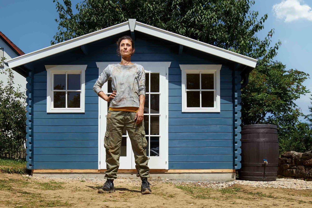 En kvinna står framför ett blått trädgårdshus med vita accenter, bredvid vilket det finns en regnvattentunna.