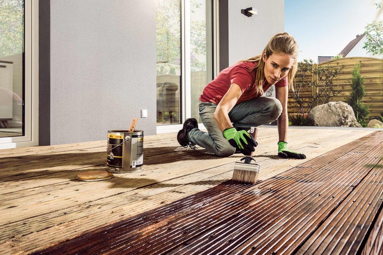 Kvinna målar en träterrass med en pensel och en burk träskyddslasyr.