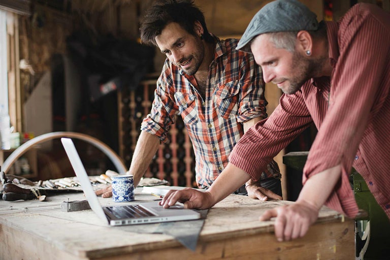 Två hantverkare arbetar på en bärbar dator i en verkstad. Verktyg och byggmaterial syns i bakgrunden.