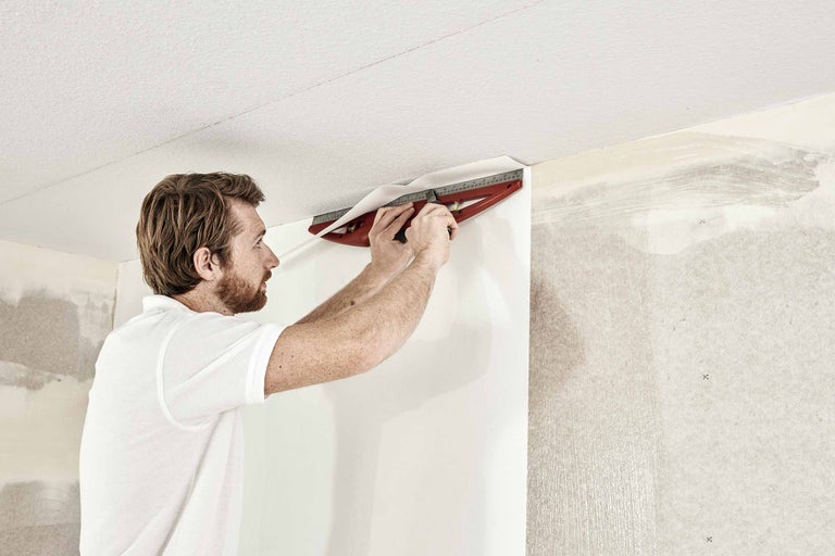 En man skär tapeter med en skärmall på en vägg.