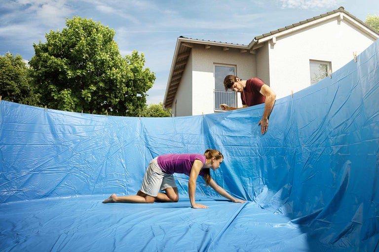 Scen med två personer som installerar en blå poolfolie, med ett bostadshus och träd i bakgrunden.