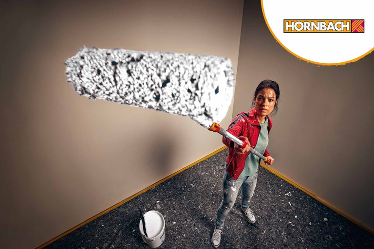 Kvinna målar en vägg med en färgroller, hink med färg står i rummet, Hornbach logotyp
