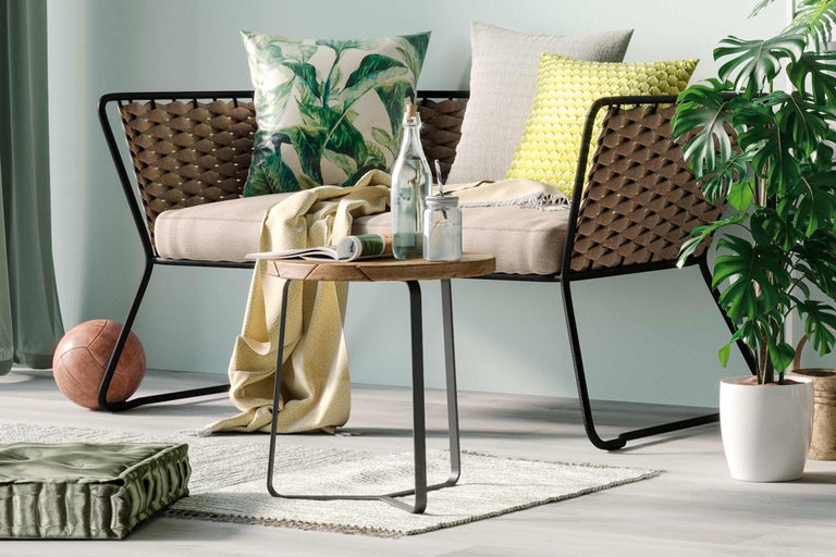 Vardagsrumsscen med rottingfåtölj, runt träbord, kuddar och krukväxt för en mysig atmosfär