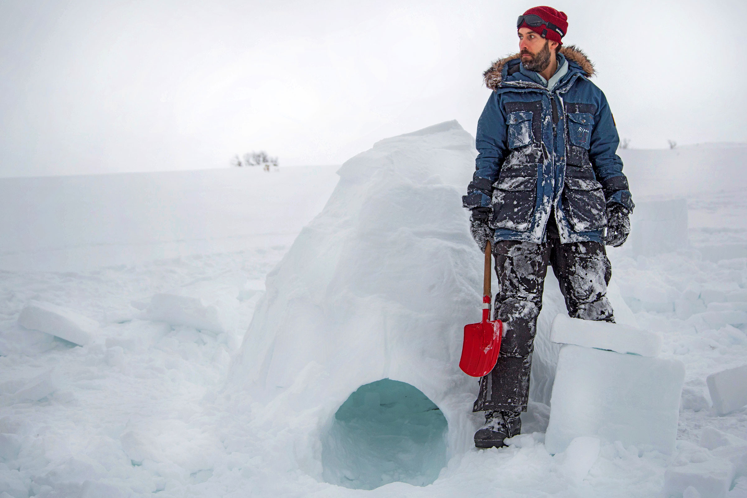 Man i vinterkläder står bredvid en igloo med en spade i snön.