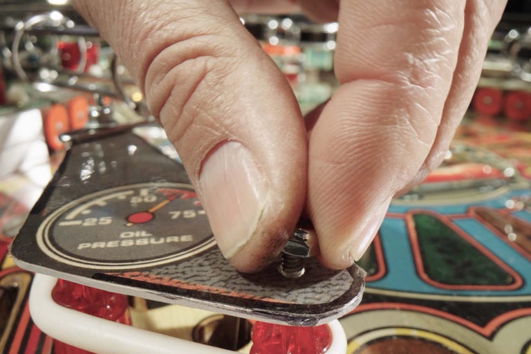 En person skruvar fast ett lock med en oljetrycksmätare på en flipperspel.