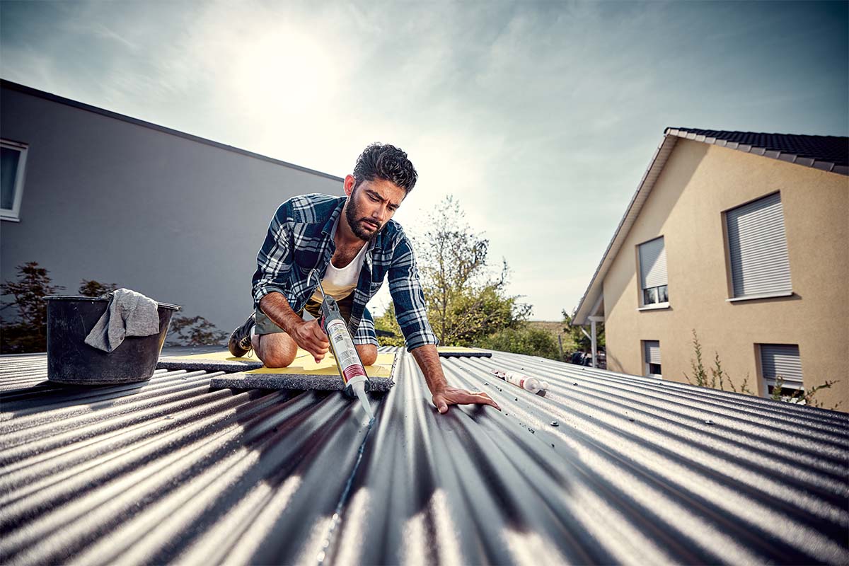 En man tätar ett metalltak med en tätningspistol. Bostadshus kan ses i bakgrunden.