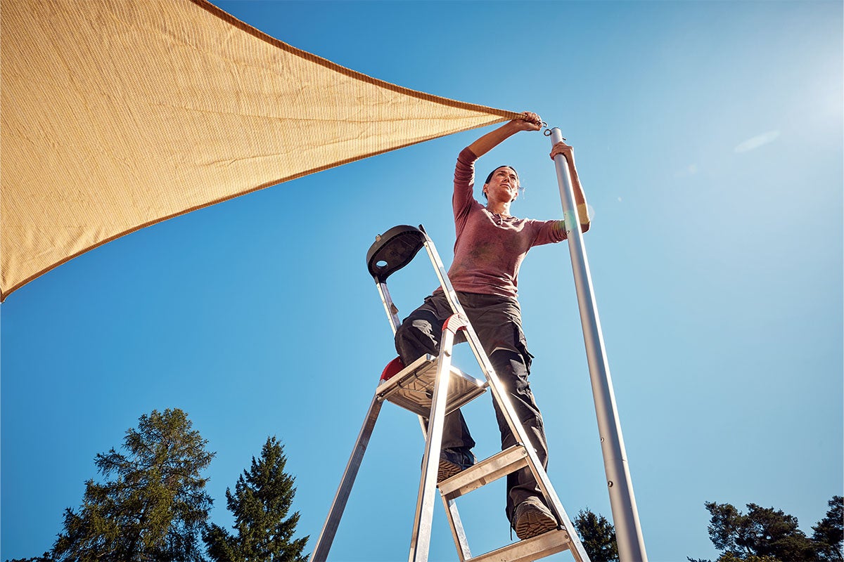 En kvinna monterar ett solsegel på en stege på en stolpe.